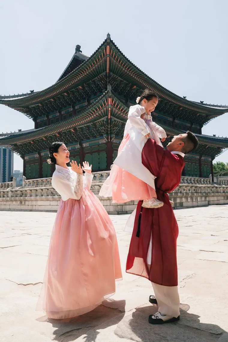 Father in crimson hanbok lifting his child while the mother cheers in the courtyard