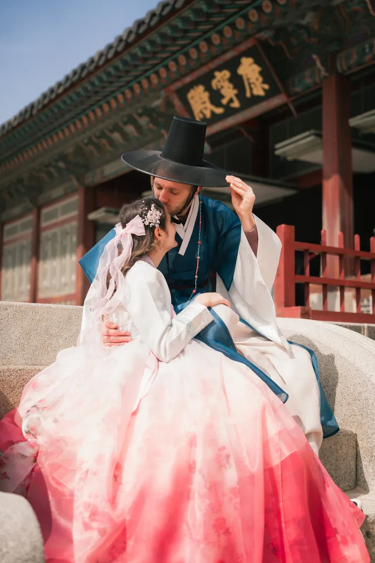 Couple in hanbok sitting close on a palace stair, the man in a gat