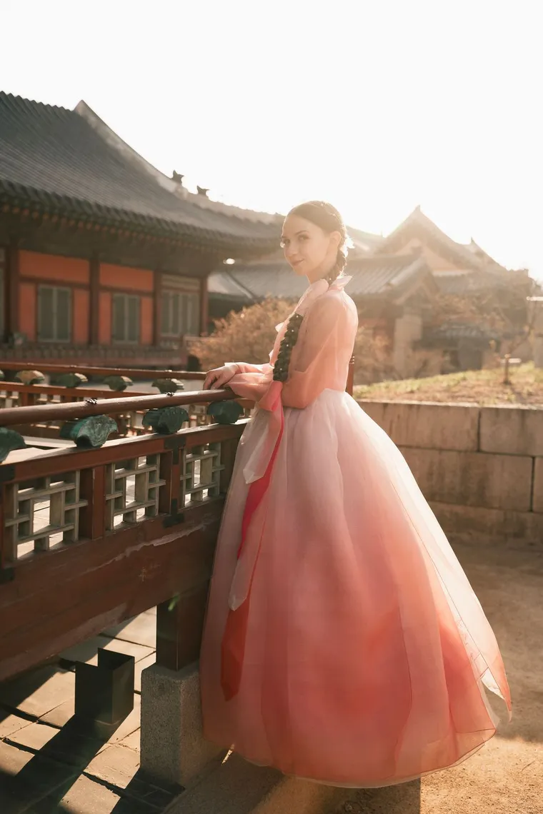 Woman in a pink hanbok at a palace railing, hanok roofs behind