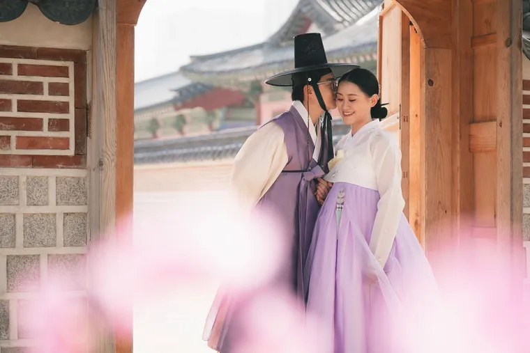 Couple in lilac hanbok leaning together beneath a palace roof