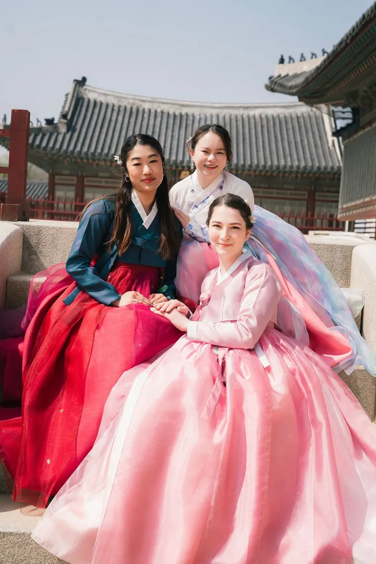 Three friends in pink and red hanbok sitting together on a palace stair
