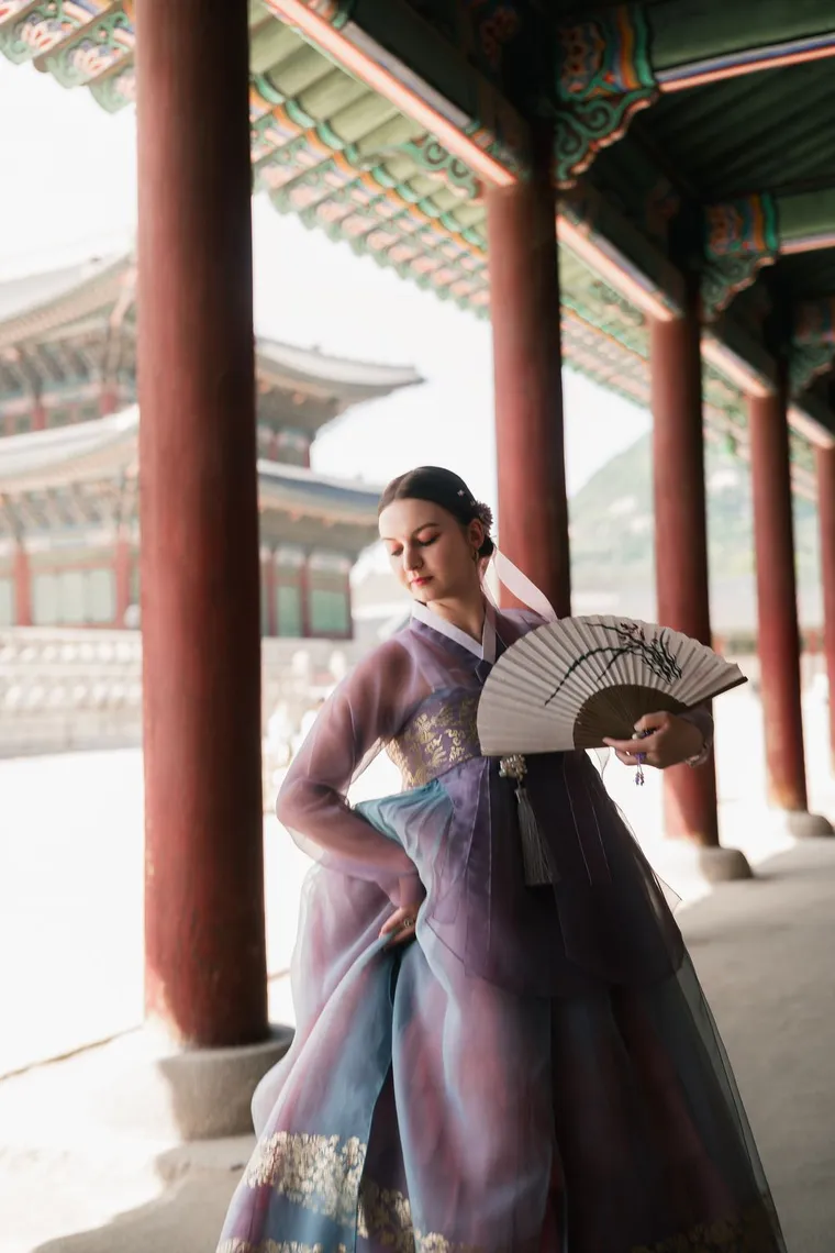 Woman in a lilac hanbok holding a fan behind her back in a colonnade