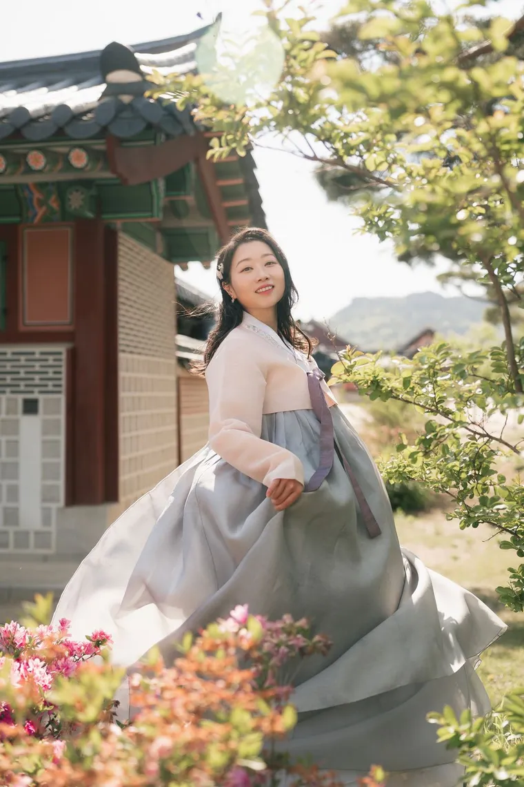 Woman in a grey and pink hanbok beside a tree in a palace garden