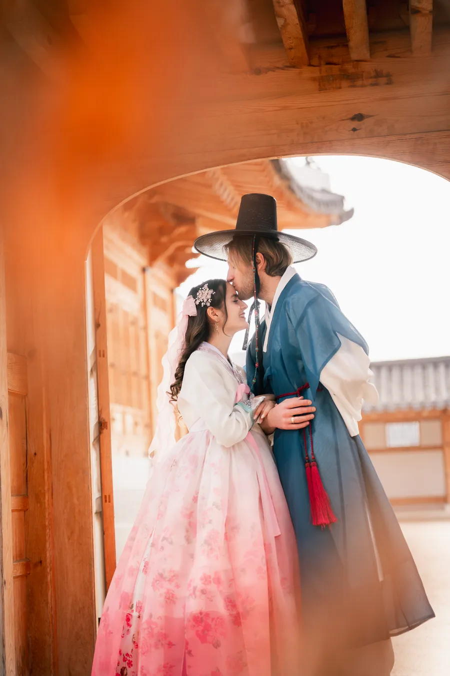 Couple in hanbok backlit in a palace corridor, morning sun flaring through the timber columns