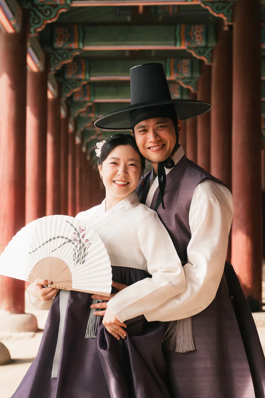 Couple in hanbok laughing together behind a folding fan in warm morning light