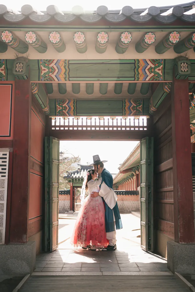 Couple in hanbok framed by an open palace gate, courtyard and rooflines behind them