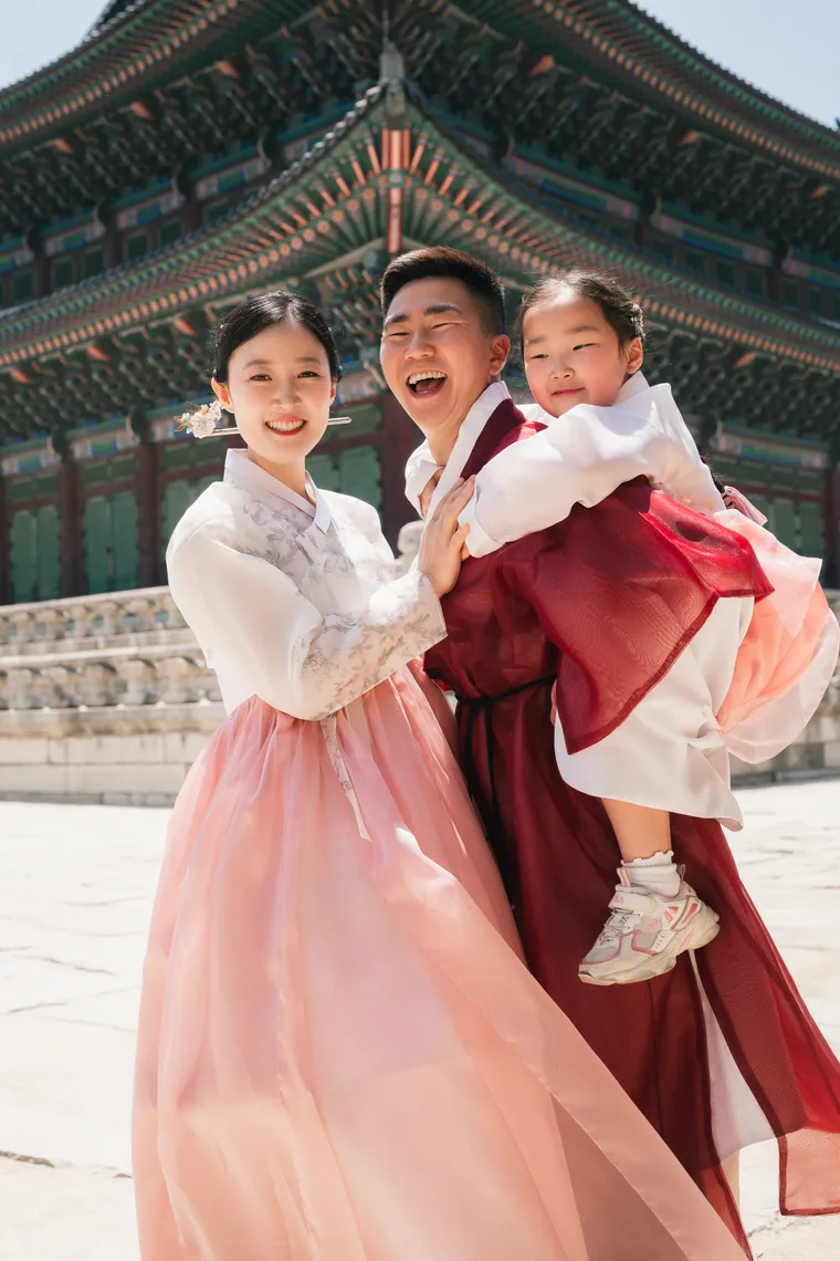 Parents lifting a child, all in hanbok, on the wide stone courtyard of Gyeongbokgung