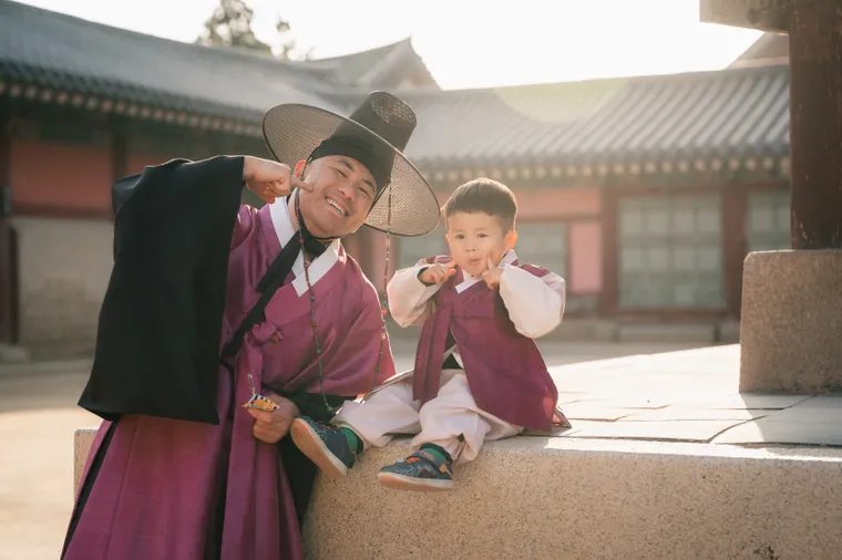 Father and small son in hanbok sitting together on palace steps, both laughing