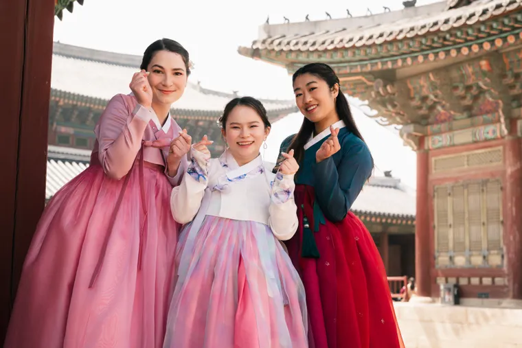 Three friends in pink, white and red hanbok making finger hearts beneath a painted palace gate
