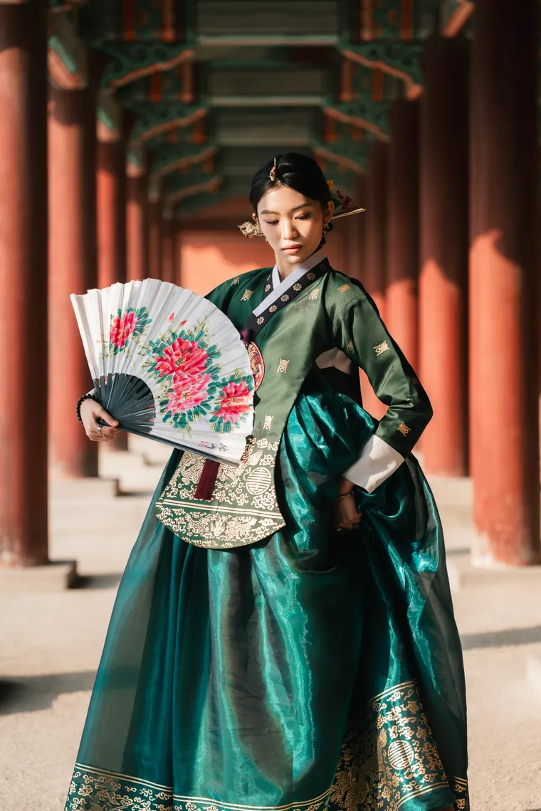 Woman in deep green silk hanbok holding a painted fan in a red-columned palace colonnade