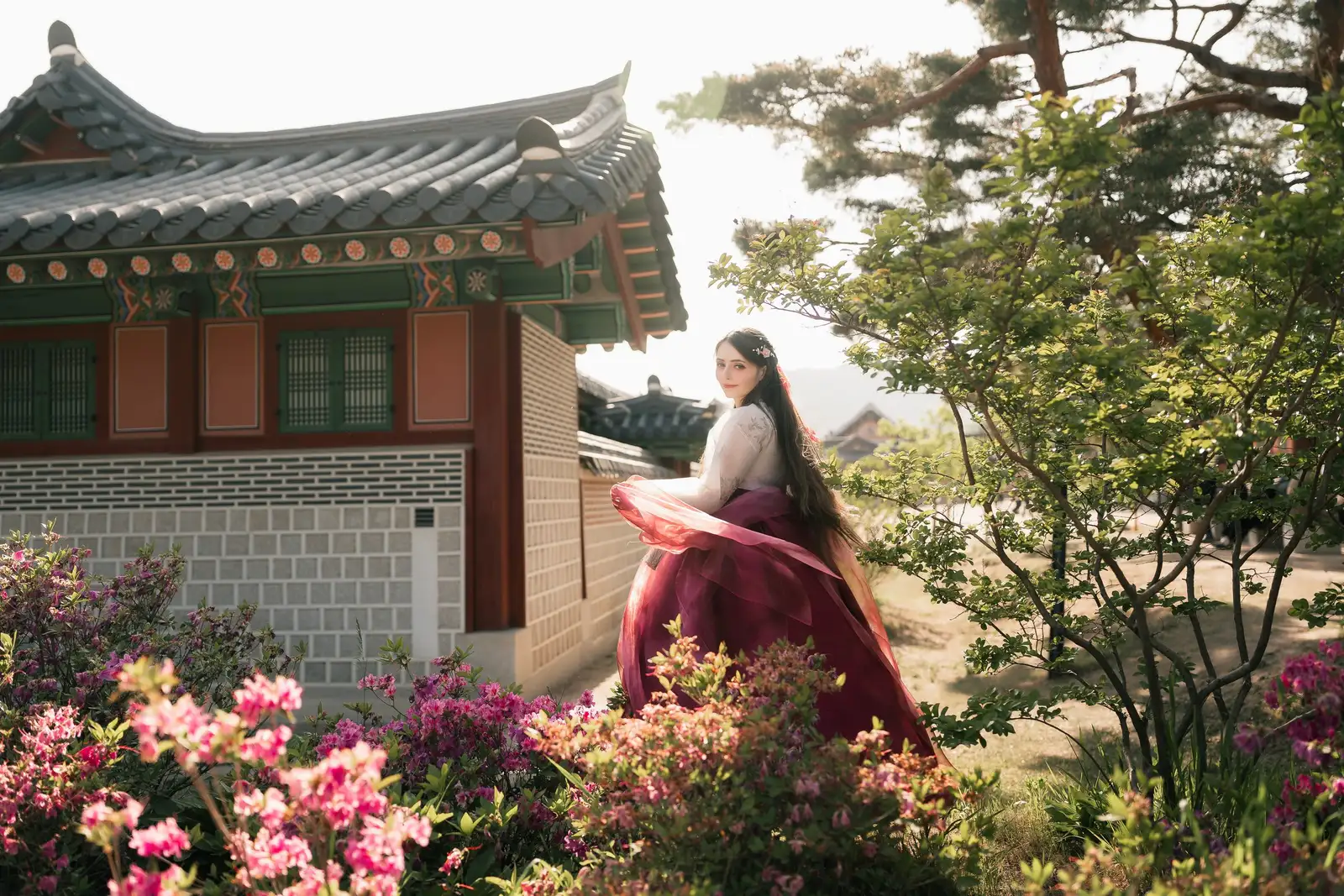 Woman in a crimson hanbok turning among azaleas beside a palace hall, backlit by evening sun
